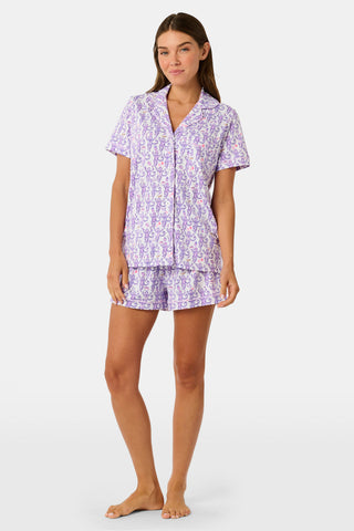 A woman is wearing the TEXPIMA Get Ready With Me Polo Pajamas — a matching short-sleeve button-up top and shorts set featuring a purple and pink patterned print — while standing against a plain light background, looking at the camera with a gentle smile.