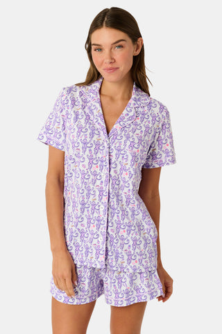A woman is wearing the TEXPIMA Get Ready With Me Polo Pajamas — a matching short-sleeve button-up top and shorts set featuring a purple and pink patterned print — while standing against a plain light background, looking at the camera with a gentle smile.