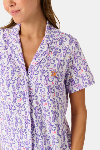 A woman is wearing the TEXPIMA Get Ready With Me Polo Pajamas — a matching short-sleeve button-up top and shorts set featuring a purple and pink patterned print — while standing against a plain light background, looking at the camera with a gentle smile.