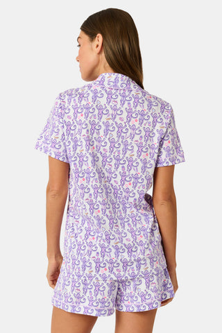 A woman is wearing the TEXPIMA Get Ready With Me Polo Pajamas — a matching short-sleeve button-up top and shorts set featuring a purple and pink patterned print — while standing against a plain light background, looking at the camera with a gentle smile.