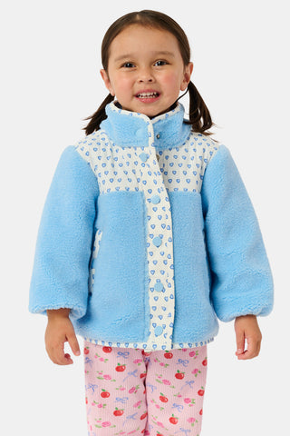 A young girl stands and smiles against a plain background, wearing the Roller Rabbit Kids Hearts Lizzie Fleece Jacket in light blue with white patterned details, paired with matching white leggings adorned with blue hearts. This cozy outerwear features soft fleece for comfort and warmth.