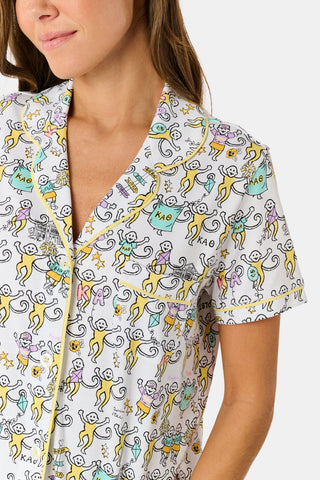 The Kappa Alpha Theta Monkey Polo Pajamas from Roller Rabbit are a short-sleeve button-up set with matching shorts, showcasing a lively print of colorful cartoon monkeys and doodles on a crisp white background. They feature an elastic waistband for comfort and are crafted from soft Pima cotton for a gentle feel.