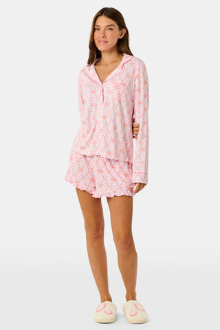 Wearing Roller Rabbit’s Lasso Bows Hayden Pajamas in pastel pink, a woman pairs the two-piece set with brown cowboy boots as she stands by a light-colored house door, smiling and resting one arm above her head while bending one leg with her foot on a step.