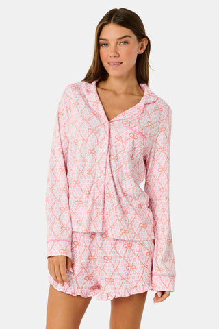 Wearing Roller Rabbit’s Lasso Bows Hayden Pajamas in pastel pink, a woman pairs the two-piece set with brown cowboy boots as she stands by a light-colored house door, smiling and resting one arm above her head while bending one leg with her foot on a step.