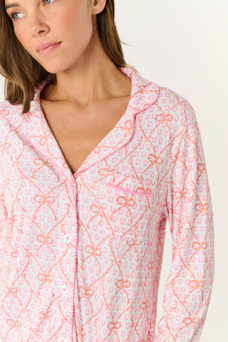 Wearing Roller Rabbit’s Lasso Bows Hayden Pajamas in pastel pink, a woman pairs the two-piece set with brown cowboy boots as she stands by a light-colored house door, smiling and resting one arm above her head while bending one leg with her foot on a step.