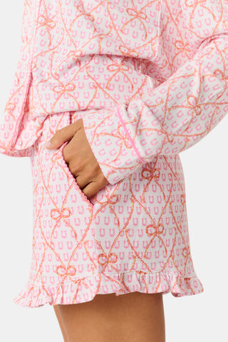Wearing Roller Rabbit’s Lasso Bows Hayden Pajamas in pastel pink, a woman pairs the two-piece set with brown cowboy boots as she stands by a light-colored house door, smiling and resting one arm above her head while bending one leg with her foot on a step.