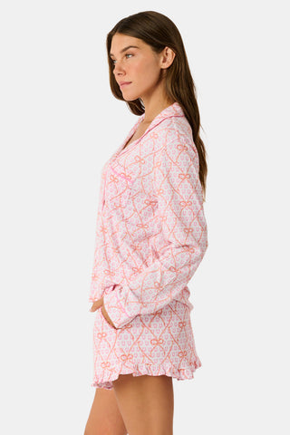 Wearing Roller Rabbit’s Lasso Bows Hayden Pajamas in pastel pink, a woman pairs the two-piece set with brown cowboy boots as she stands by a light-colored house door, smiling and resting one arm above her head while bending one leg with her foot on a step.
