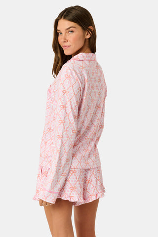 Wearing Roller Rabbit’s Lasso Bows Hayden Pajamas in pastel pink, a woman pairs the two-piece set with brown cowboy boots as she stands by a light-colored house door, smiling and resting one arm above her head while bending one leg with her foot on a step.