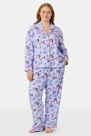 A young child with curly hair stands barefoot against a plain white background, wearing Roller Rabbit’s Kids London Monkey Pajamas—light blue long-sleeve pajamas adorned with playful, colorful patterns featuring guards, clocks, and iconic British landmarks.