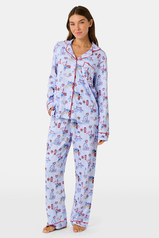 A young child with curly hair stands barefoot against a plain white background, wearing Roller Rabbit’s Kids London Monkey Pajamas—light blue long-sleeve pajamas adorned with playful, colorful patterns featuring guards, clocks, and iconic British landmarks.