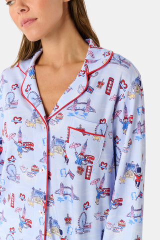 A young child with curly hair stands barefoot against a plain white background, wearing Roller Rabbit’s Kids London Monkey Pajamas—light blue long-sleeve pajamas adorned with playful, colorful patterns featuring guards, clocks, and iconic British landmarks.