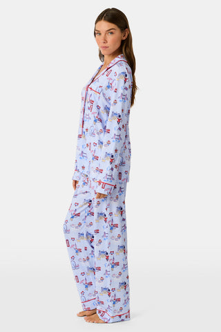 A young child with curly hair stands barefoot against a plain white background, wearing Roller Rabbit’s Kids London Monkey Pajamas—light blue long-sleeve pajamas adorned with playful, colorful patterns featuring guards, clocks, and iconic British landmarks.