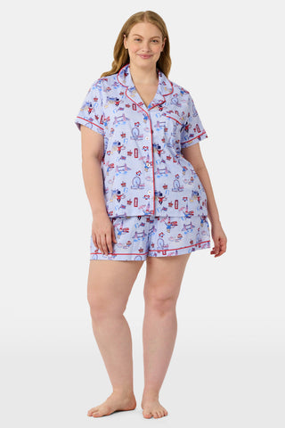 A woman stands and smiles against a plain white background, wearing the Roller Rabbit London Monkey Polo Pajamas—a light blue Pima cotton set featuring cartoon prints and red trim.