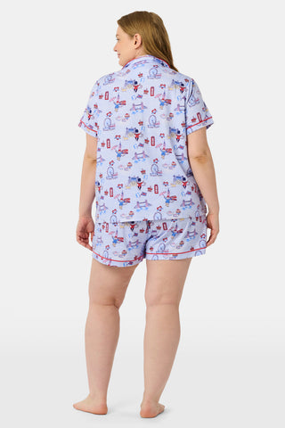 A woman stands and smiles against a plain white background, wearing the Roller Rabbit London Monkey Polo Pajamas—a light blue Pima cotton set featuring cartoon prints and red trim.
