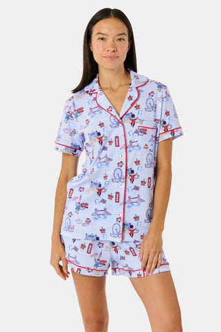 A young child with curly hair stands barefoot against a plain white background, wearing Roller Rabbit’s Kids London Monkey Pajamas—light blue long-sleeve pajamas adorned with playful, colorful patterns featuring guards, clocks, and iconic British landmarks.