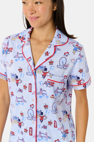 A young child with curly hair stands barefoot against a plain white background, wearing Roller Rabbit’s Kids London Monkey Pajamas—light blue long-sleeve pajamas adorned with playful, colorful patterns featuring guards, clocks, and iconic British landmarks.