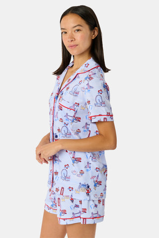 A young child with curly hair stands barefoot against a plain white background, wearing Roller Rabbit’s Kids London Monkey Pajamas—light blue long-sleeve pajamas adorned with playful, colorful patterns featuring guards, clocks, and iconic British landmarks.