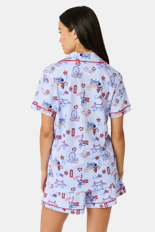 A woman stands and smiles against a plain white background, wearing the Roller Rabbit London Monkey Polo Pajamas—a light blue Pima cotton set featuring cartoon prints and red trim.
