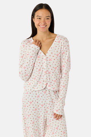 A woman wearing the Roller Rabbit Mini Monkey Blossom Pointelle Allie V-Neck Cardigan—a long-sleeve, white pajama top featuring a small pink floral print and crafted from soft Pima cotton—stands against a plain white background, smiling gently.