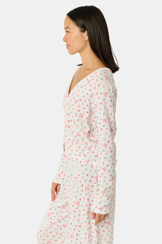 A woman wearing the Roller Rabbit Mini Monkey Blossom Pointelle Allie V-Neck Cardigan—a long-sleeve, white pajama top featuring a small pink floral print and crafted from soft Pima cotton—stands against a plain white background, smiling gently.