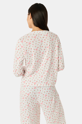 A woman wearing the Roller Rabbit Mini Monkey Blossom Pointelle Allie V-Neck Cardigan—a long-sleeve, white pajama top featuring a small pink floral print and crafted from soft Pima cotton—stands against a plain white background, smiling gently.