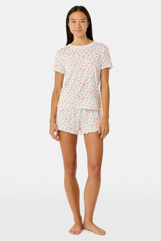 A smiling woman with curly hair stands outdoors near a window, wearing the Roller Rabbit Mini Monkey Blossom Pointelle Classic Tee—a white short-sleeve top featuring a delicate pink floral print.