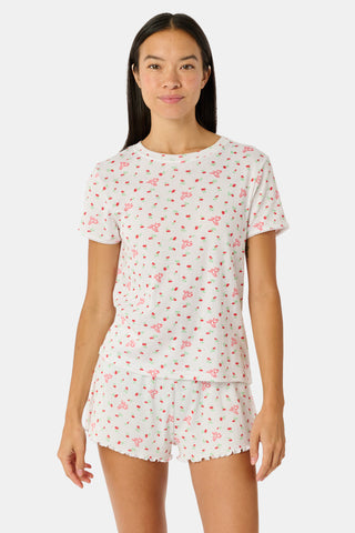 A smiling woman with curly hair stands outdoors near a window, wearing the Roller Rabbit Mini Monkey Blossom Pointelle Classic Tee—a white short-sleeve top featuring a delicate pink floral print.