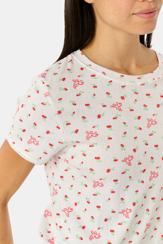 A smiling woman with curly hair stands outdoors near a window, wearing the Roller Rabbit Mini Monkey Blossom Pointelle Classic Tee—a white short-sleeve top featuring a delicate pink floral print.