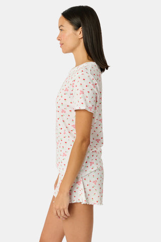 A smiling woman with curly hair stands outdoors near a window, wearing the Roller Rabbit Mini Monkey Blossom Pointelle Classic Tee—a white short-sleeve top featuring a delicate pink floral print.
