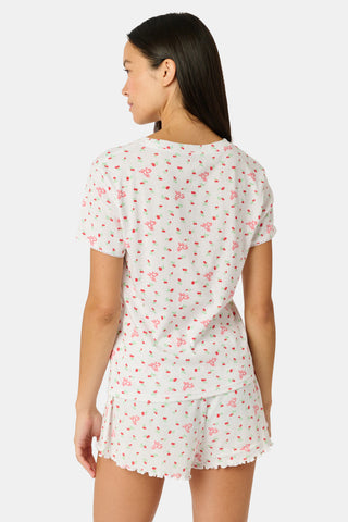 A smiling woman with curly hair stands outdoors near a window, wearing the Roller Rabbit Mini Monkey Blossom Pointelle Classic Tee—a white short-sleeve top featuring a delicate pink floral print.
