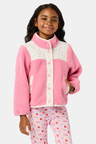 A young girl smiles, wearing the Roller Rabbit Kids Mini Monkey Blossom Lizzie Fleece Jacket, featuring white patterned panels, and matching pink leggings with colorful fruit prints, standing against a plain light gray background.