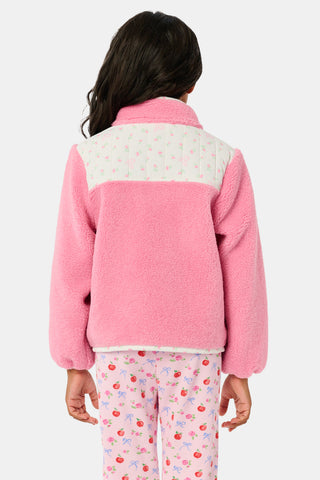 A young girl smiles, wearing the Roller Rabbit Kids Mini Monkey Blossom Lizzie Fleece Jacket, featuring white patterned panels, and matching pink leggings with colorful fruit prints, standing against a plain light gray background.