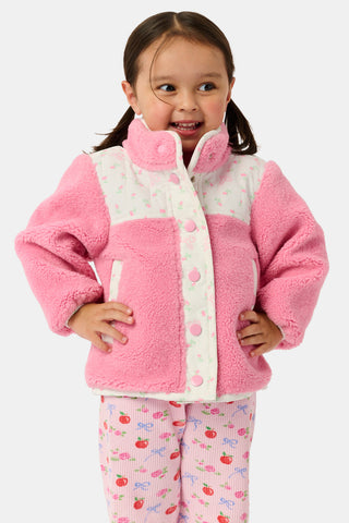 A young girl smiles, wearing the Roller Rabbit Kids Mini Monkey Blossom Lizzie Fleece Jacket, featuring white patterned panels, and matching pink leggings with colorful fruit prints, standing against a plain light gray background.