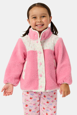A young girl smiles, wearing the Roller Rabbit Kids Mini Monkey Blossom Lizzie Fleece Jacket, featuring white patterned panels, and matching pink leggings with colorful fruit prints, standing against a plain light gray background.