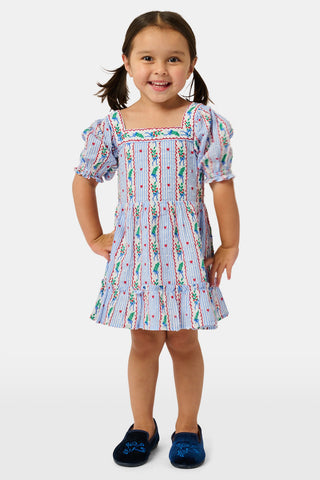A girl wears the SANSKRITI Kids Mistletoe Stripe Lottie Dress in a blue and white-striped cotton print with puff sleeves, a square neckline, and red heart accents, paired with blue velvet Mary Jane flats.