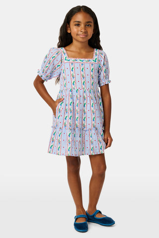 A girl wears the SANSKRITI Kids Mistletoe Stripe Lottie Dress in a blue and white-striped cotton print with puff sleeves, a square neckline, and red heart accents, paired with blue velvet Mary Jane flats.