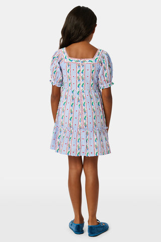 A girl wears the SANSKRITI Kids Mistletoe Stripe Lottie Dress in a blue and white-striped cotton print with puff sleeves, a square neckline, and red heart accents, paired with blue velvet Mary Jane flats.