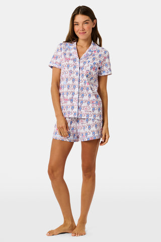 The Roller Rabbit Pi Beta Phi Monkey Polo Pajamas feature a short-sleeved button-up top and matching shorts, adorned with a playful pink and blue monkey print on a white background. Crafted from soft Pima cotton with an elastic waistband for comfort, these pajamas are made in Peru.