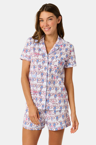 The Roller Rabbit Pi Beta Phi Monkey Polo Pajamas feature a short-sleeved button-up top and matching shorts, adorned with a playful pink and blue monkey print on a white background. Crafted from soft Pima cotton with an elastic waistband for comfort, these pajamas are made in Peru.