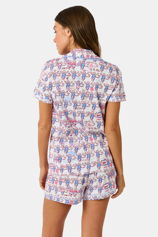The Roller Rabbit Pi Beta Phi Monkey Polo Pajamas feature a short-sleeved button-up top and matching shorts, adorned with a playful pink and blue monkey print on a white background. Crafted from soft Pima cotton with an elastic waistband for comfort, these pajamas are made in Peru.