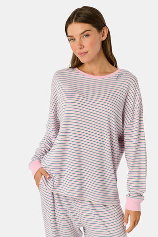 A woman wearing the Roller Rabbit Striped Brynn Oversized Tee—an ultra-comfy loungewear top with thin pink, blue, and white horizontal stripes and pink cuffs and collar—stands with one hand in her pocket against a plain background.