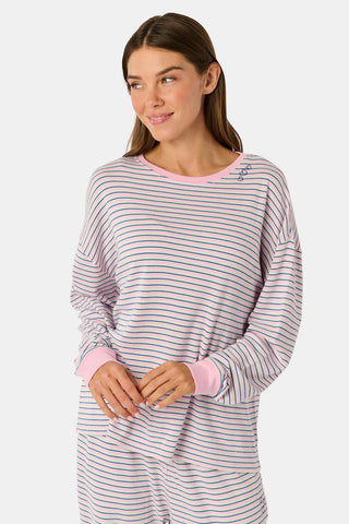 A woman wearing the Roller Rabbit Striped Brynn Oversized Tee—an ultra-comfy loungewear top with thin pink, blue, and white horizontal stripes and pink cuffs and collar—stands with one hand in her pocket against a plain background.