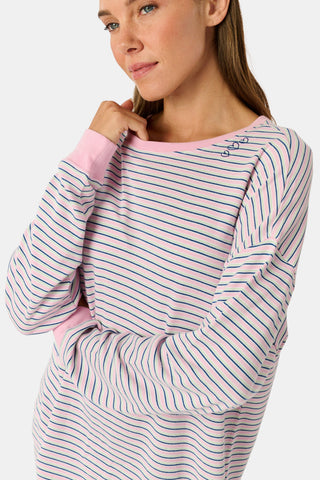 A woman wearing the Roller Rabbit Striped Brynn Oversized Tee—an ultra-comfy loungewear top with thin pink, blue, and white horizontal stripes and pink cuffs and collar—stands with one hand in her pocket against a plain background.