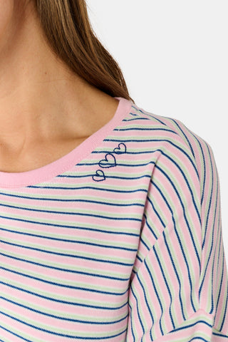 A woman wearing the Roller Rabbit Striped Brynn Oversized Tee—an ultra-comfy loungewear top with thin pink, blue, and white horizontal stripes and pink cuffs and collar—stands with one hand in her pocket against a plain background.