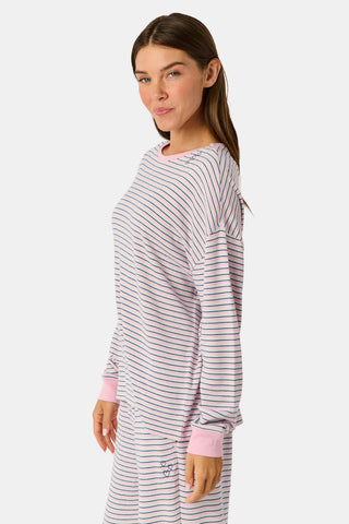 A woman wearing the Roller Rabbit Striped Brynn Oversized Tee—an ultra-comfy loungewear top with thin pink, blue, and white horizontal stripes and pink cuffs and collar—stands with one hand in her pocket against a plain background.