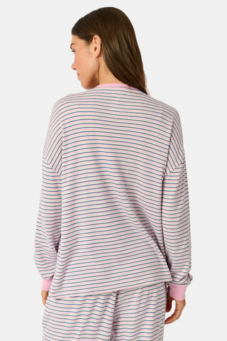 A woman wearing the Roller Rabbit Striped Brynn Oversized Tee—an ultra-comfy loungewear top with thin pink, blue, and white horizontal stripes and pink cuffs and collar—stands with one hand in her pocket against a plain background.