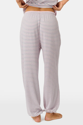 A person stands barefoot on a white background wearing the Roller Rabbit Striped Eva Pant—loose, light-colored pima cotton pajama pants featuring thin blue and red horizontal stripes. Their top is lifted slightly, revealing the elastic waistband.