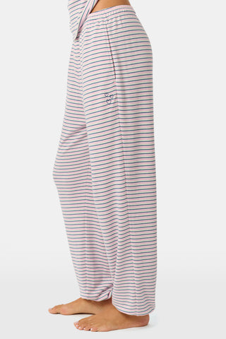 A person stands barefoot on a white background wearing the Roller Rabbit Striped Eva Pant—loose, light-colored pima cotton pajama pants featuring thin blue and red horizontal stripes. Their top is lifted slightly, revealing the elastic waistband.