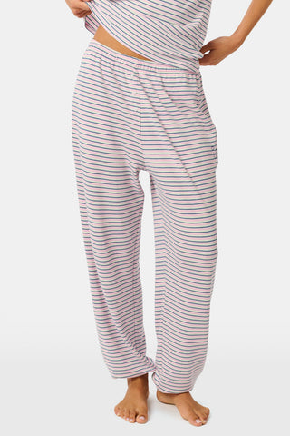 A person stands barefoot on a white background wearing the Roller Rabbit Striped Eva Pant—loose, light-colored pima cotton pajama pants featuring thin blue and red horizontal stripes. Their top is lifted slightly, revealing the elastic waistband.