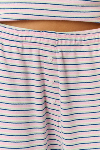 A person stands barefoot on a white background wearing the Roller Rabbit Striped Eva Pant—loose, light-colored pima cotton pajama pants featuring thin blue and red horizontal stripes. Their top is lifted slightly, revealing the elastic waistband.